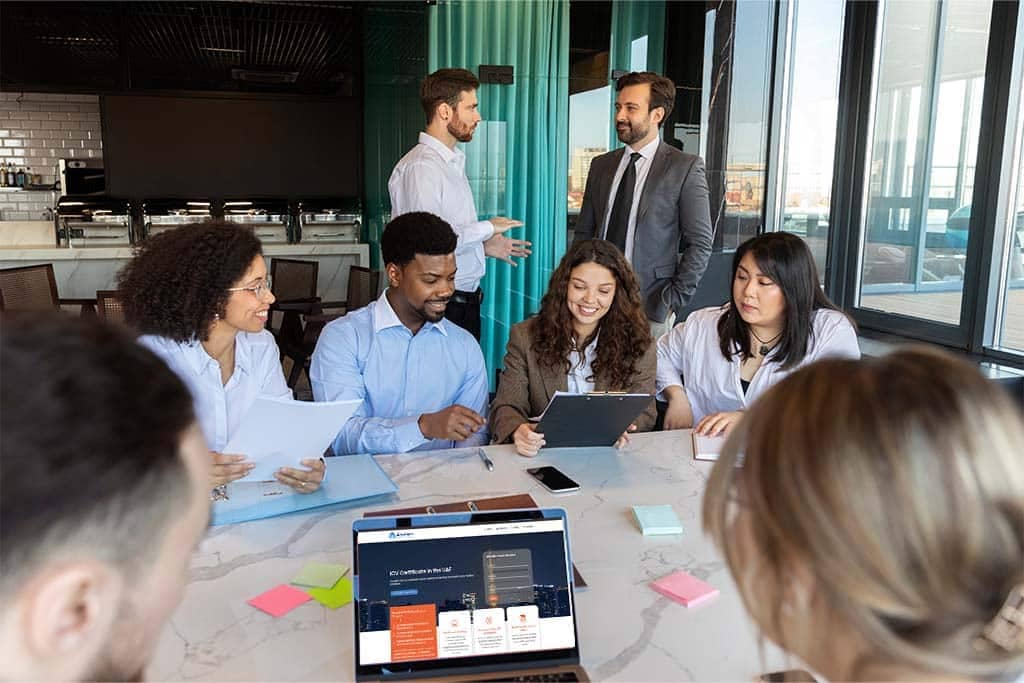 A group of seven people are in a modern office setting. Four are seated at a table engaging with documents and a tablet, while three are standing in the background, talking. There is a laptop on the table displaying an ICV Certificate webpage. Bright windows offer a city view.