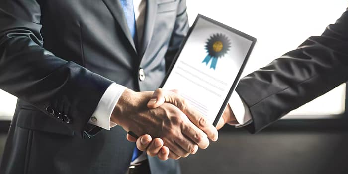A few individuals are gathered indoors, each holding a tablet. They are dressed in formal attire, including blazers and ties, indicative of a business environment. This setting suggests they may be engaged in a professional discussion or presentation related to their work as consultants.