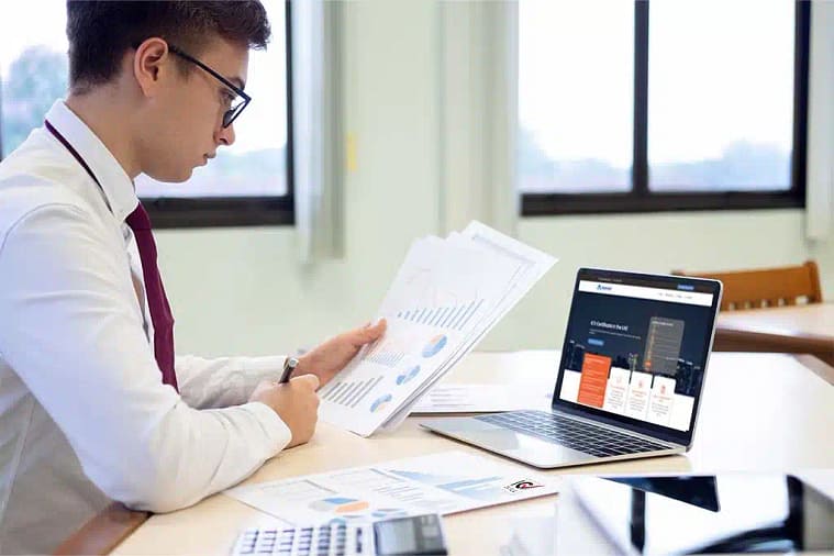 A person wearing a white shirt and maroon tie sits at a desk, reviewing printed charts and graphs, including an ICV Certificate for UAE Business. They have open a laptop displaying graphs and text. The desk also has a calculator and additional documents spread out. The room has large windows with light streaming in.