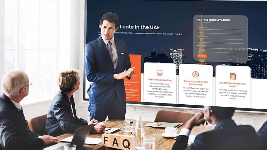 A man in a business suit is giving a presentation to three seated colleagues. They are in a conference room, looking at a large screen displaying a website titled "Abstract" with information on VAT services in the UAE. A "Get Free Consultation" form is visible on the screen.